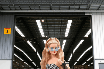 A blonde Barbie in a black-and-white dress standing inside a large warehouse.