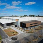 An aerial view of a large, modern DHL Health Logistics warehouse under a blue sky.