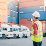 Worker in hard hat and safety vest using a radio at a shipping yard.