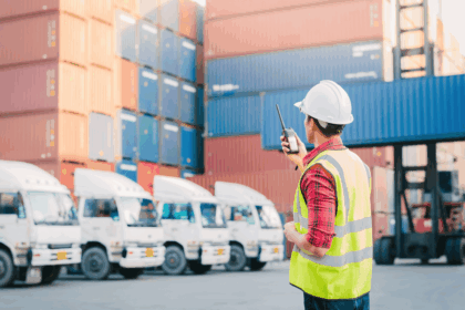 Worker in hard hat and safety vest using a radio at a shipping yard.