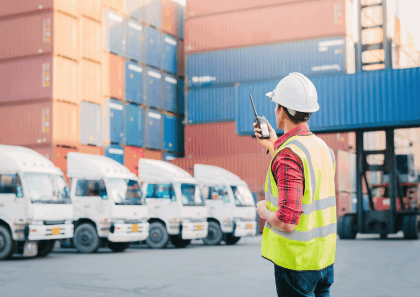 Worker in hard hat and safety vest using a radio at a shipping yard.