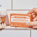 A hand holding a bar of Dr. Bronner's soap next to a soapy brush.
