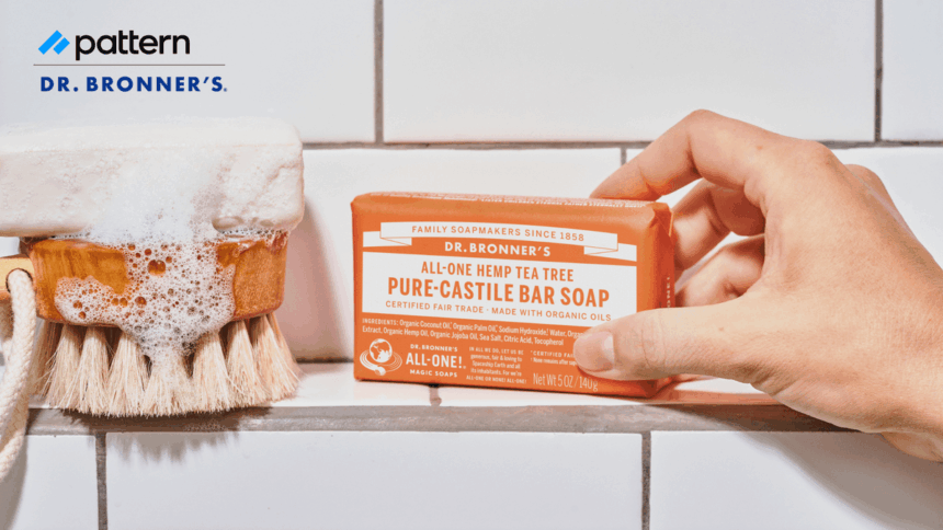 A hand holding a bar of Dr. Bronner's soap next to a soapy brush.