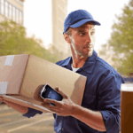 Delivery person in uniform holding a package and a handheld scanner outdoors.