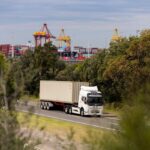 An electric semi-truck hauling a shipping container on a road near a port.