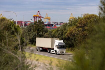 An electric semi-truck hauling a shipping container on a road near a port.