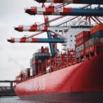 A large Hamburg Süd container ship docked under towering red and white cranes.