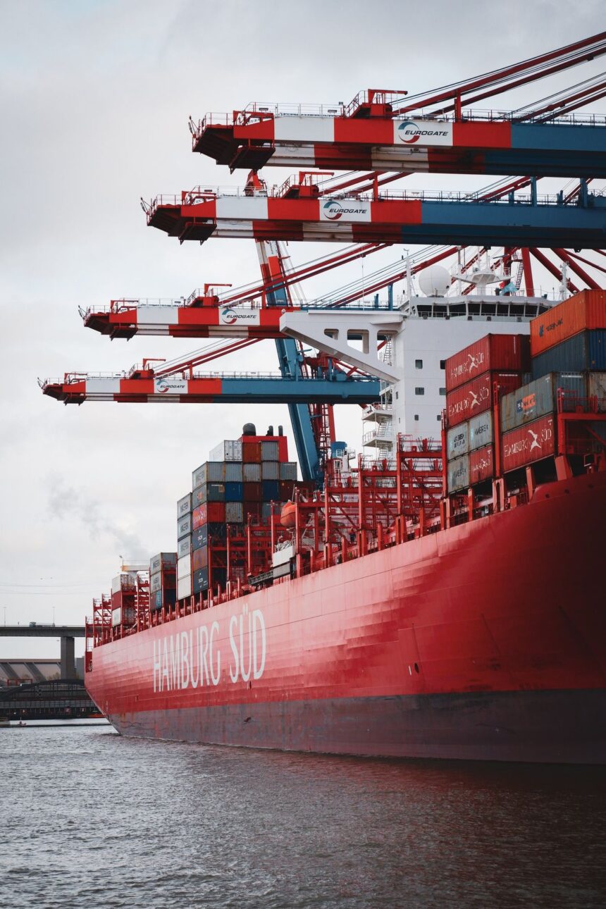 A large Hamburg Süd container ship docked under towering red and white cranes.