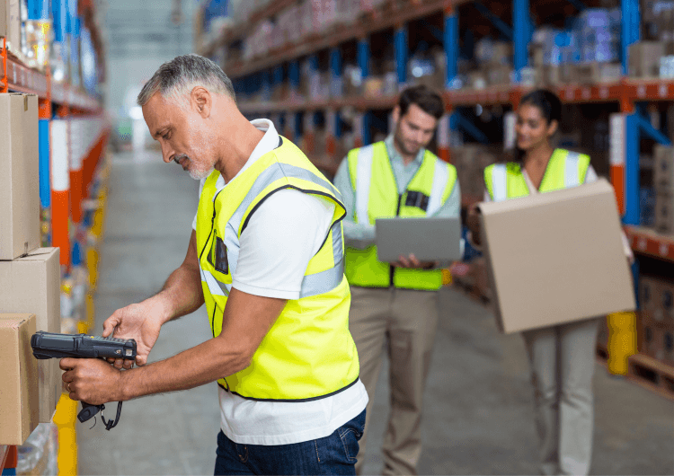 Warehouse workers wearing safety vests scanning items and carrying boxes in a storage facility.
