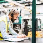Bearded man wearing a high-visibility vest and headset, working on a laptop in a factory.