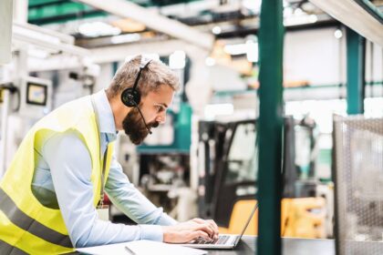Bearded man wearing a high-visibility vest and headset, working on a laptop in a factory.