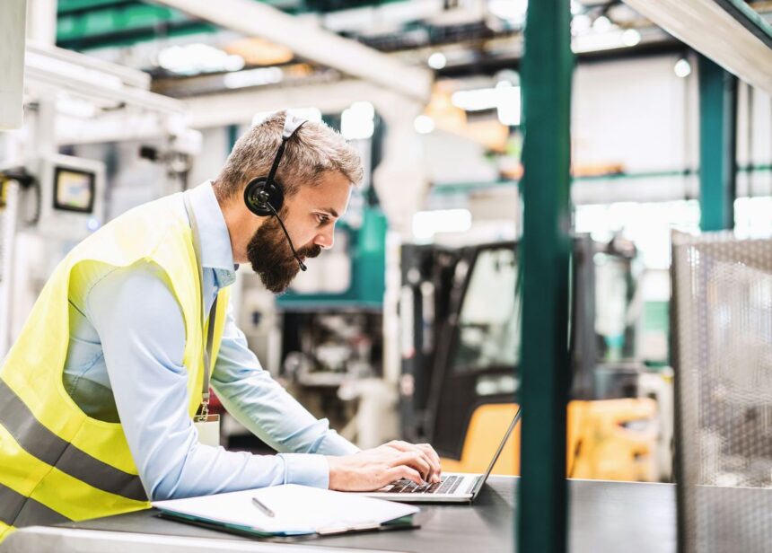 Bearded man wearing a high-visibility vest and headset, working on a laptop in a factory.