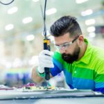Factory worker in safety glasses uses an electric screwdriver on an electronic circuit board.