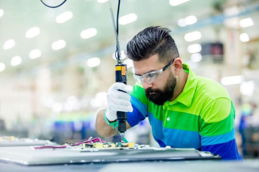 Factory worker in safety glasses uses an electric screwdriver on an electronic circuit board.
