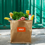 A brown paper bag labeled VOLY filled with fresh groceries sits on the ground.