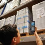 Warehouse worker using a handheld scanner to check inventory on high storage shelves.