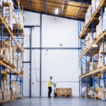 A warehouse worker operates a pallet jack between tall rows of stored cardboard boxes.