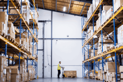 A warehouse worker operates a pallet jack between tall rows of stored cardboard boxes.
