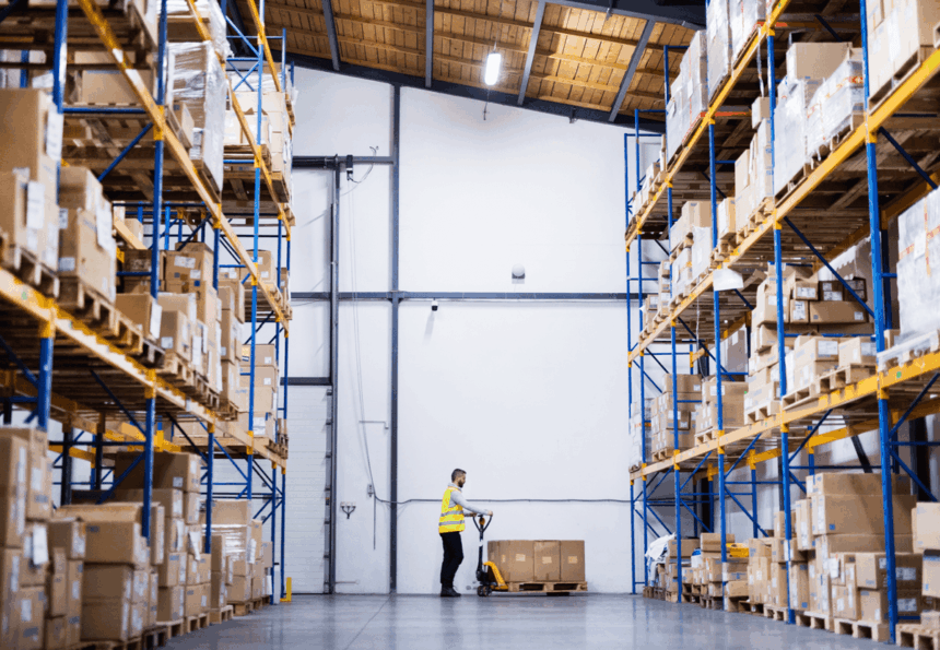 A warehouse worker operates a pallet jack between tall rows of stored cardboard boxes.