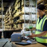 Warehouse worker in safety vest scanning items at a workstation in a large warehouse.