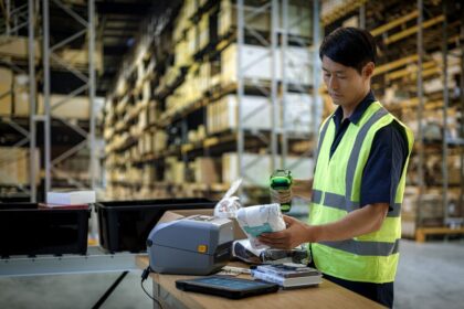 Warehouse worker in safety vest scanning items at a workstation in a large warehouse.