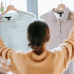 A person in a yellow sweater comparing two shirts hanging on hangers.