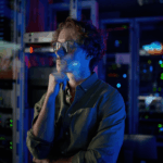 A thoughtful man wearing glasses stands in a server room with glowing lights.