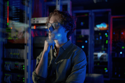 A thoughtful man wearing glasses stands in a server room with glowing lights.