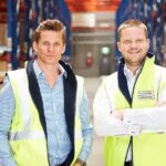 Two men in high-visibility vests standing in a warehouse with storage racking.