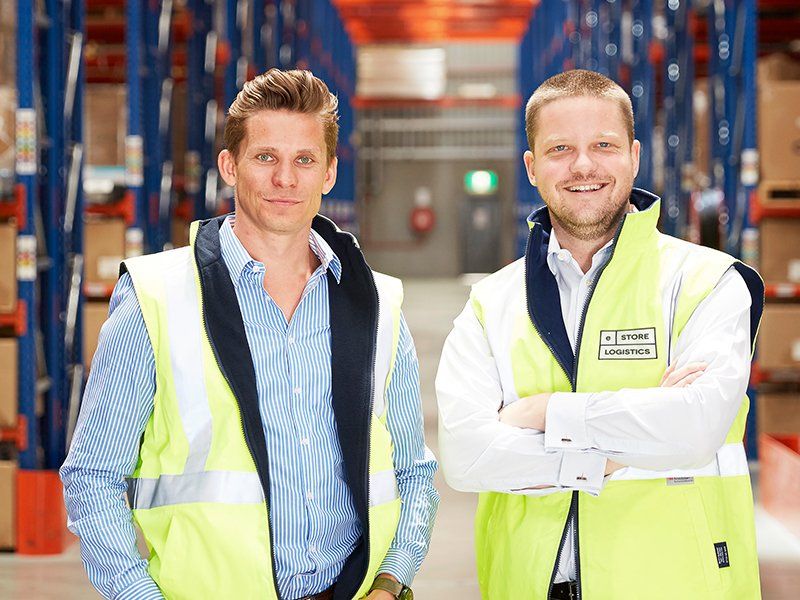 Two men in high-visibility vests standing in a warehouse with storage racking.