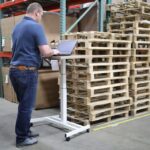 A man uses a laptop on a standing desk in a warehouse.