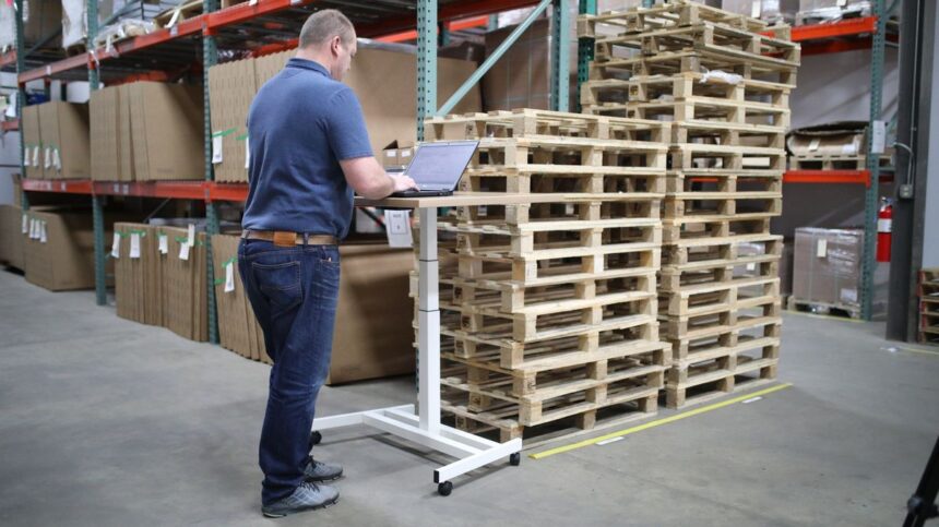A man uses a laptop on a standing desk in a warehouse.