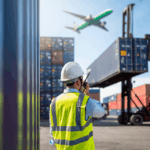 A logistics worker in a safety vest coordinates near shipping containers as a plane flies.