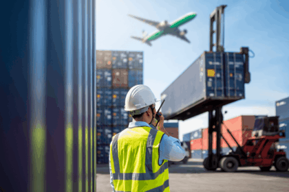A logistics worker in a safety vest coordinates near shipping containers as a plane flies.