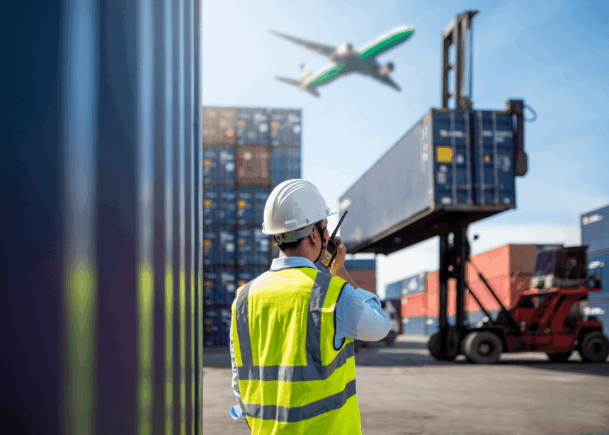 A logistics worker in a safety vest coordinates near shipping containers as a plane flies.