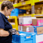 Warehouse worker using a wrist-mounted scanner to process a blue plastic crate.