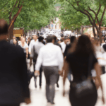 Crowds of people walking down a sunny, tree-lined city street.