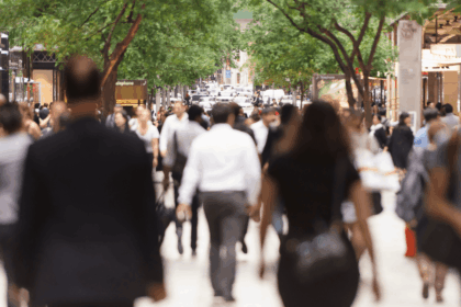 Crowds of people walking down a sunny, tree-lined city street.