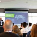 A speaker presents a global logistics events map to an audience in a conference room.