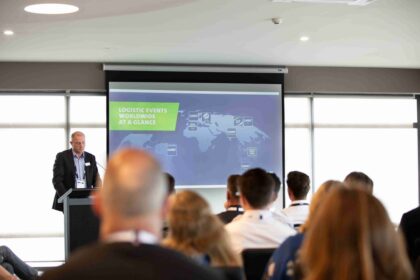 A speaker presents a global logistics events map to an audience in a conference room.