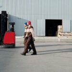 Two warehouse workers in uniform walking past a parked forklift outside a factory.