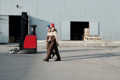 Two warehouse workers in uniform walking past a parked forklift outside a factory.