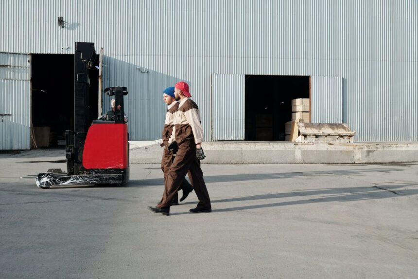 Two warehouse workers in uniform walking past a parked forklift outside a factory.
