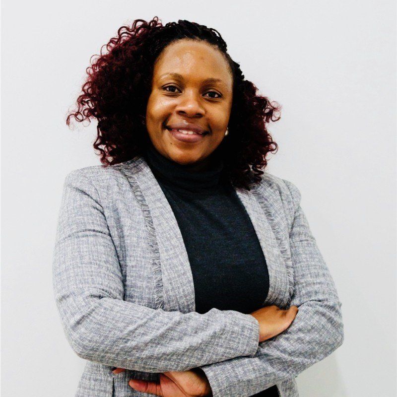 Smiling woman with curly auburn hair, wearing a gray blazer, arms crossed against white.