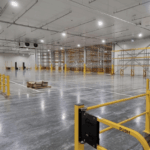 Large, modern, empty warehouse with concrete floors, yellow safety railings, and high industrial racking.