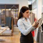 A shop assistant uses a tablet while checking clothing on a retail store rack.
