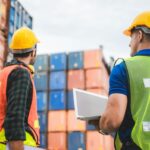 Two workers in safety vests and helmets stand before stacked shipping containers with a laptop.