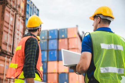Two workers in safety vests and helmets stand before stacked shipping containers with a laptop.