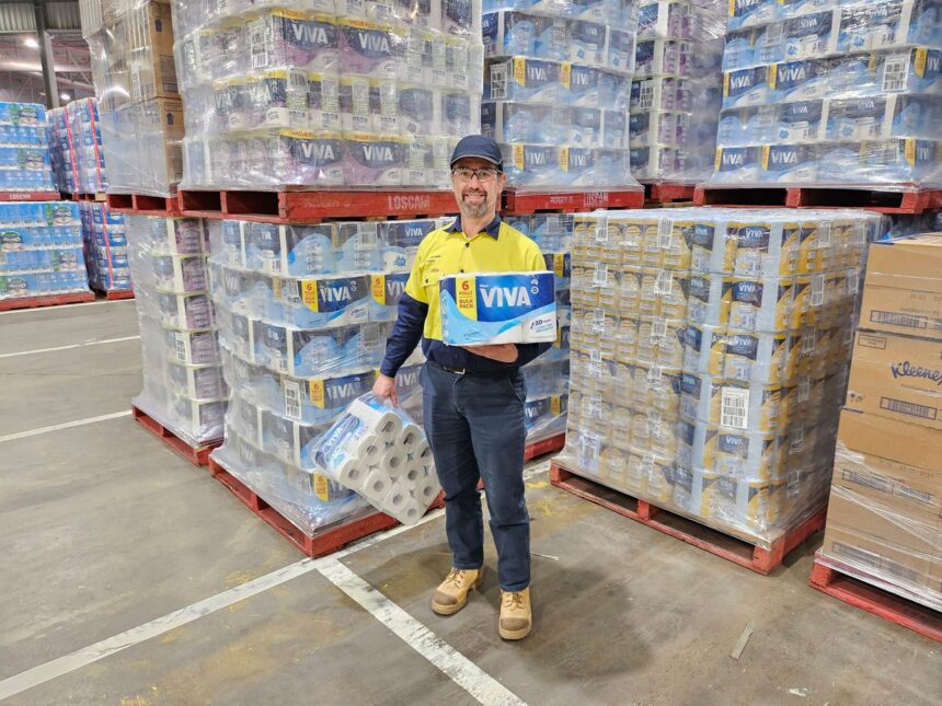 A warehouse worker standing between pallets of Viva paper towels holding two packages.