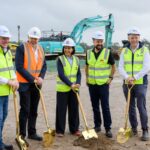 Five people wearing hard hats and high-vis vests hold ceremonial shovels at a construction site.